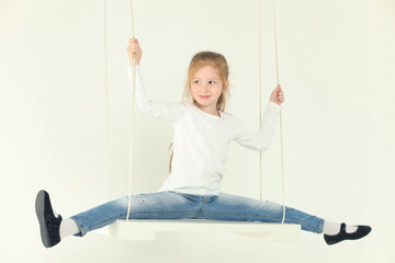Happy little girl in jeans swings with leg-split in white studio and looks away