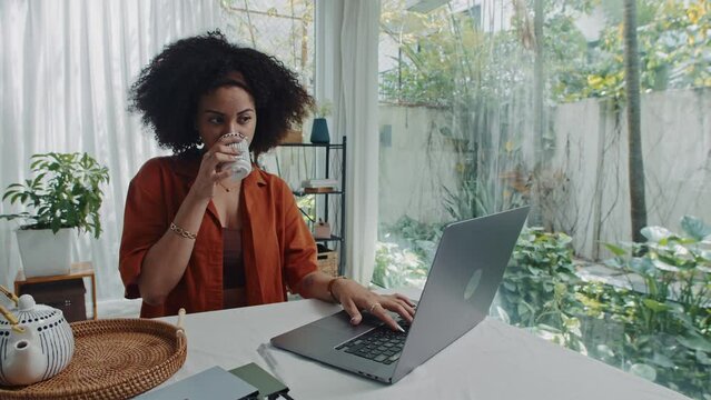 Medium shot of female remote worker filling form on laptop and drinking tea sitting at wireless laptop
