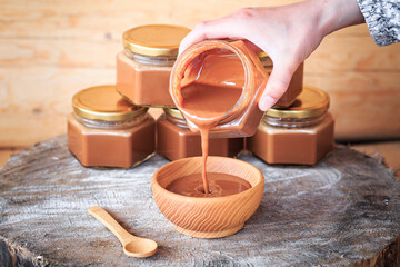 A girl's hands pour boiled condensed milk into a wooden plate, close-up against the background of glass jars filled with condensed milk, food photography, wooden background. Vertical orientation.