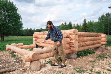 A man with a beard builds a wooden house from logs with an ax in nature, logs in the foreground, construction, village, summer, greenery, forest, bathhouse, carpenters, peasants.