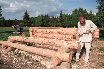 Two men are building a wooden house from logs in nature, cutting logs with axes, logs in the foreground, construction, village, summer, greenery, forest, bathhouse, carpenters.