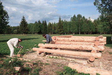 Two men are building a wooden house from logs in nature, cutting logs with axes, logs in the foreground, construction, village, summer, greenery, forest, summer, bathhouse, carpenters.