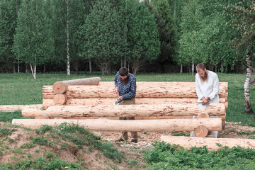 Two men are building a wooden house from logs in nature, cutting logs with axes, logs in the foreground, construction, village, summer, greenery, forest, summer.
