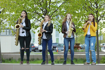 Four young pretty women in jeans play saxophone outdoor at spring day