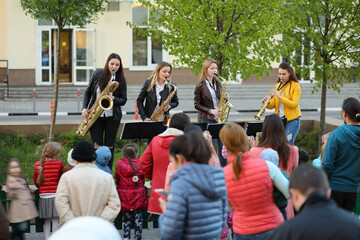 Four young women in jeans play saxophone outdoor at spring day, people listen their