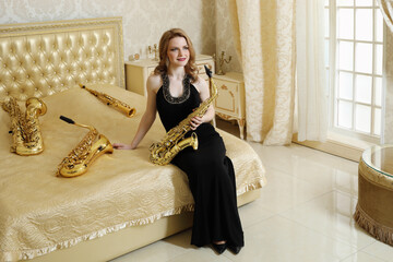 Pretty woman in black dress sits with sax on bed with wind instruments in baroque studio - obrazy, fototapety, plakaty