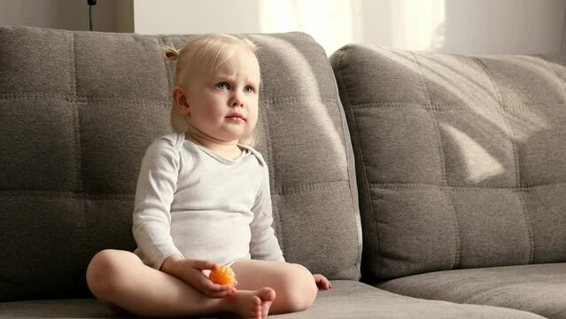 A little girl with blond hair sits on a gray sofa and watches cartoons on the tv. Child watching tv. Daylighting.