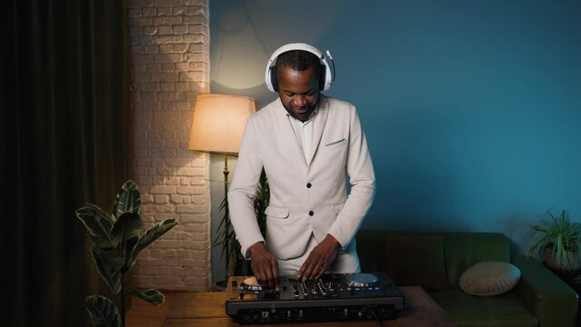 The DJ is playing a set at a house party. The man is dressed in a white suit and white headphones.