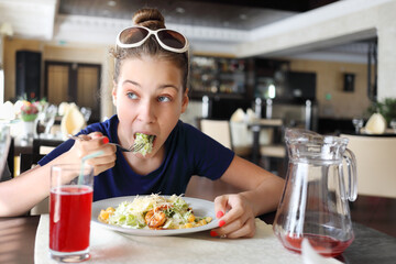 Pretty hungry girl with sunglasses eats salad in cafe, juice is on table
