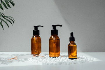 Amber bottles with facial treatment on a white little decorative stones with green leaves on backdrop. Front view