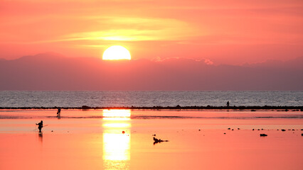 Stunning pink and orange sunset reflecting over ocean water with silhouetted people fishing at low...