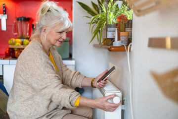 Senior woman holding mobile phone while adjusting the thermostat on the radiator at home - 762350212