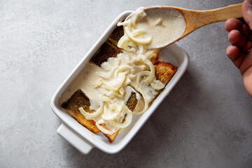 Baking dish with roasted whitefish fillet on kitchen counter. Hand adding creamy sauce to fish with spoon.