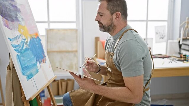 A thoughtful man in an apron contemplating a canvas in a bright art studio.