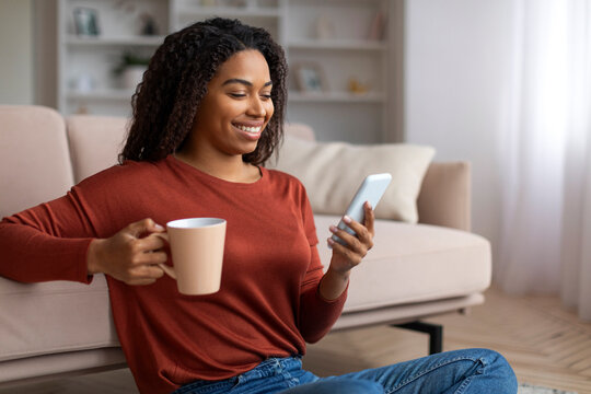 Domestic Leisure. Smiling Black Woman Relaxing On Floor With Smartphone And Coffee