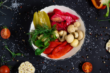 Assorted pickled vegetables. Canned tomatoes, cucumbers, chili peppers in a round black plate on a dark background top view. Appetizer of canned vegetables. Home preservation.