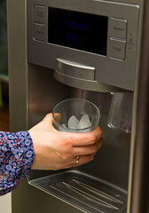 Ice making fridge detail with a female hand holding a glass of ice cubes.