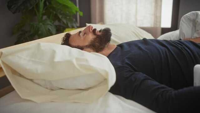 Bearded man waking up in a cozy bedroom, stretching and smiling contentedly.