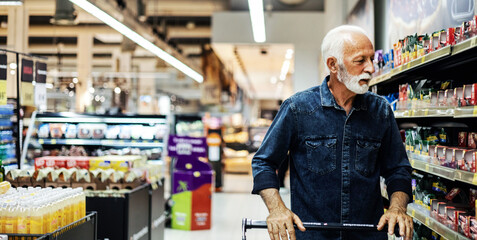 Mature Caucasian man is shopping in a supermarket, leaning against his cart with a smile on his...