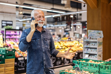 Senior Caucasian man is having a phone call while shopping in a supermarket. Elderly man is standing in a supermarket isle and having a conversation over the phone with a shopping cart in front of him