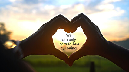 Inspirational quote - We can only learn to love by loving. On person making heart shape for love sign with fingers against the colorful sunset sunrise light background. Romantic love quotes concept. - Powered by Adobe