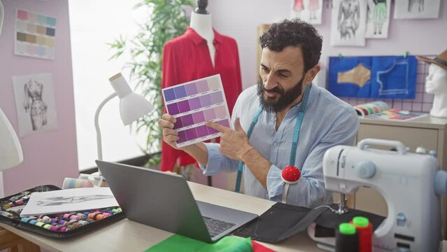 A bearded tailor in a studio examines fabric swatches and a fashion sketch on a laptop with a sewing machine and mannequin nearby.