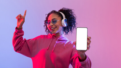 Energetic black lady dancing with headphones, holding smartphone with blank screen, vibrant backdrop