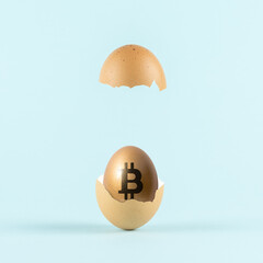Golden egg with a bitcoin sign in cracked eggshell against pastel blue background. Minimal investment concept.