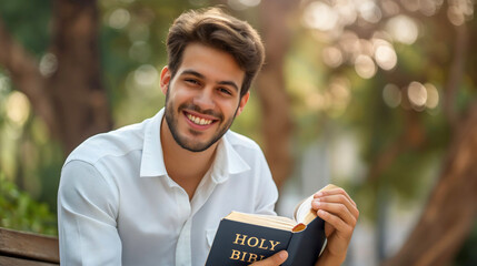 Handsome young man wearing white shirt, smiling at the camera, holding the Holy Bible Christian scripture book in his hands, sitting on a wooden bench in a park on a sunny summer day, studying reading