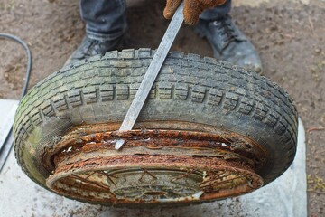 a worker in brown mittens uses tools to remove an old black rubber tire from a rusty iron rim from a motorcycle wheel during the day on the street