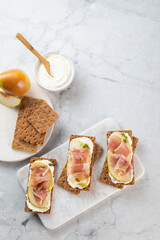 Ideal snack wholewheat crispbread with ricotta cheese, prosciutto, sliced pear and thyme on light grey marble cutting board. Healthy snack concept. Italian cuisine