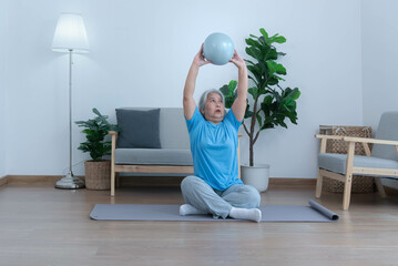 Asian elderly woman exercising at home by stretching her arm muscles and using a ball as an exercise aid for retirement people and health care concept.