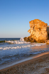 Vertical seascape. A stone cliff on the seashore with a sandy beach is washed by a wave.