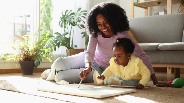 young african american mother teaching her baby to paint on the floor sitting