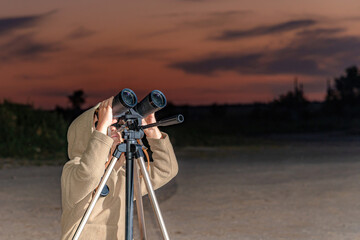 A boy looks through binoculars on a tripod against the background of an evening sunset.