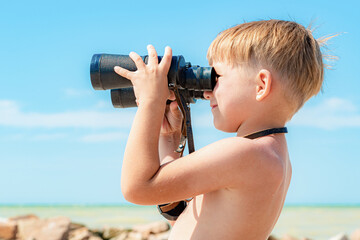 A boy with binoculars looks at the sea against a blue sky.