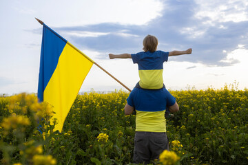 dad and son, sitting on shoulders, arms outstretched, stand with the flag of Ukraine among blooming...