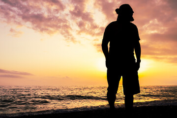 A man stands on a beach at sunset, looking out at the ocean