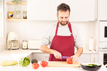 A man is cutting tomatoes on a cutting board in a kitchen. He is wearing a red apron and a grey shirt. There are several other vegetables on the counter, including a cucumber and a carrot