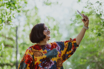 Happy woman taking a selfie with her mobile phone in a blooming park. Young entrepreneur with digital devices capturing a selfie, staying connected in an urban green space