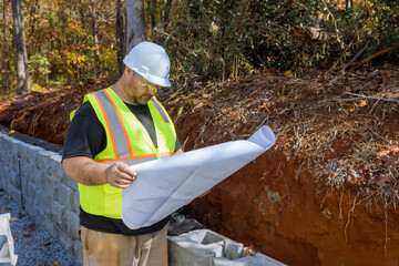 Construction worker studies used by blueprint plan when mounting retaining wall by cement blocks are being