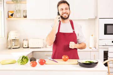 A man is talking on his cell phone while standing in a kitchen. He is wearing a red apron and is smiling. There are several vegetables on the counter, including tomatoes, lettuce, and carrots