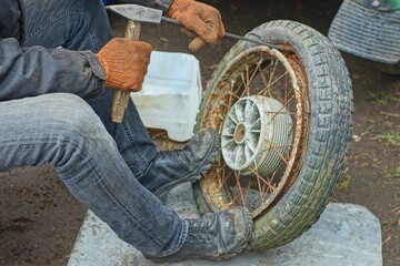  a worker in brown mittens removes an old black rubber tire with a hammer from a rusty iron rim...
