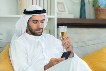 Portrait of a smiling Arabian man holding a glass of champagne and using mobile phone at home