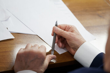 An elderly man in a business suit sits at a desk with papers and holds a fountain pen. Writing a...