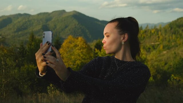 Young woman making selfies using mobile phone standing on top of mountain peak. Happy girl taking photos on cellphone camera, scenic landscape view on background