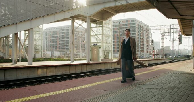 A woman in formal clothes and a coat smiles as she walks along the station platform with a suitcase in her hand