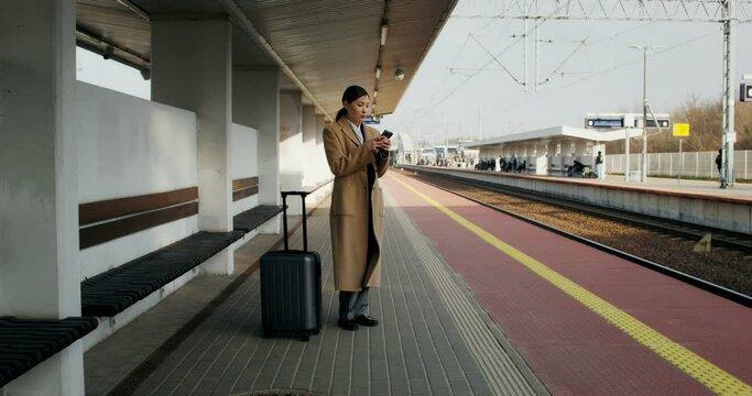 A woman in formal clothes and a coat uses a mobile phone standing on the platform of a railway station. Her luggage is nearby