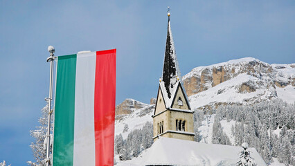 Church with Italian flag in Arabba, Italy (province Belluno). Dolomites mountains in the background with a blue sky and lots of snow, after heavy snowfall. 