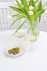 Table set with two glasses of white wine, vase with white tulips, plate with olives on white table on chair and bricks background.